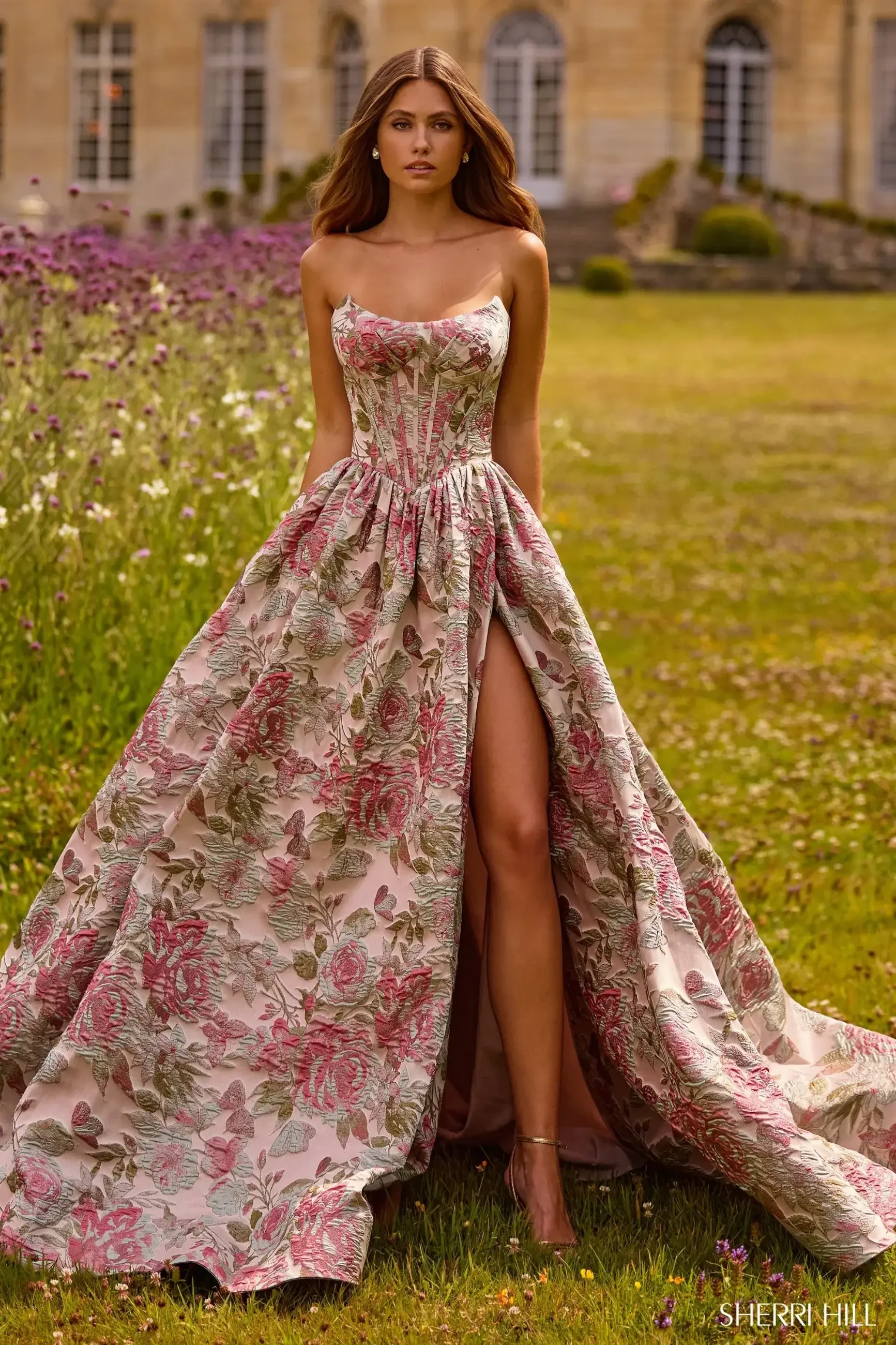 A woman wearing a floral gown with a high slit, standing in a garden with flowers and an elegant building in the background.